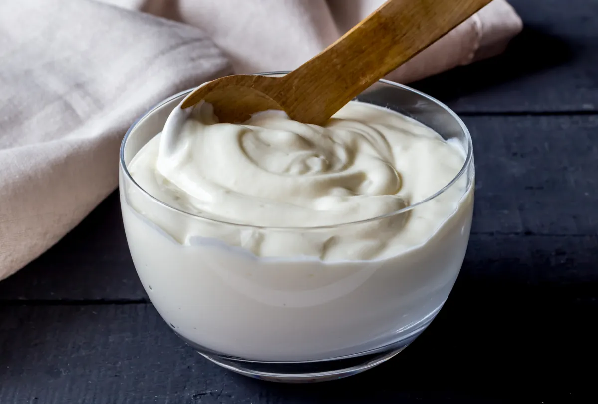 Clear bowl of white yogurt with a wooden spoon on a dark surface.