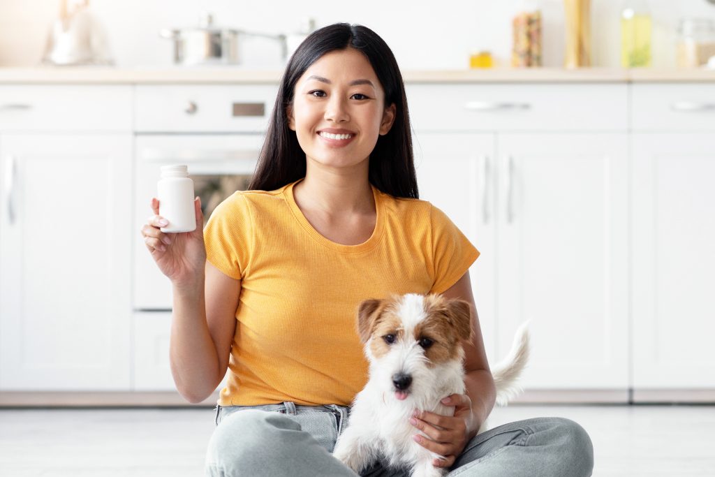 94416678 cheerful asian lady with cute doggy showing pets supplement jar